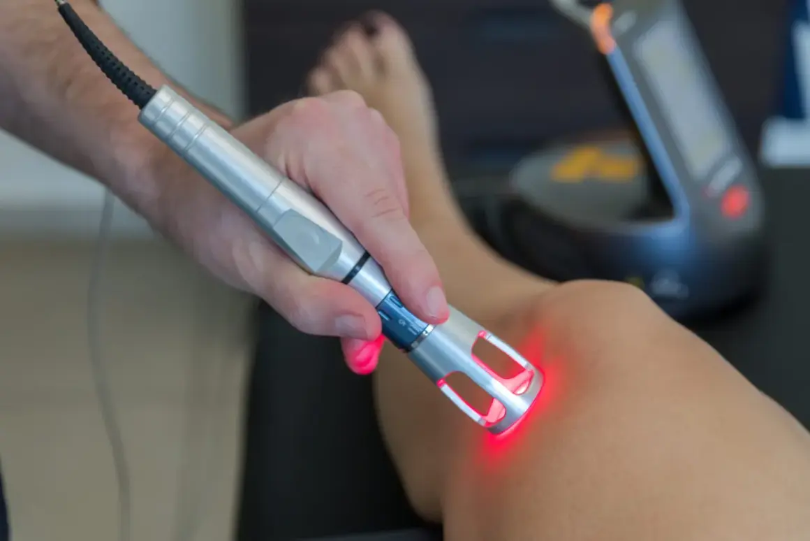 Dłoń trzymająca urządzenie z czerwonym światłem, które emituje laser wysokoenergetyczny na skórę nogi.
