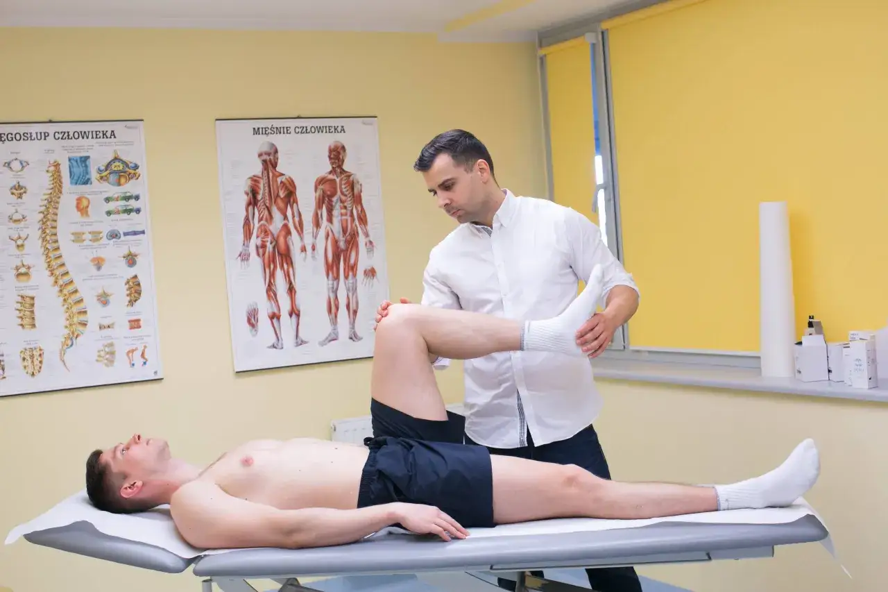Fizjoterapeuta bada nogę pacjenta, podejrzewając naderwany mięsień. Na ścianie wiszą anatomiczne plakaty.