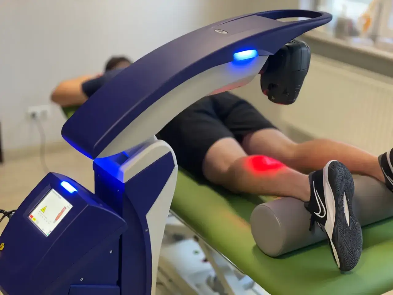 Pacjent leży na kozetce, a na jego kolanie działa nowoczesny laser wysokoenergetyczny, emitując czerwone światło.