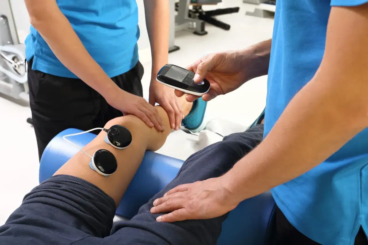 Fizjoterapeuta stosuje elektrody EMS do nogi pacjenta, kontrolując urządzenie. To część terapii, która pomaga w regeneracji.