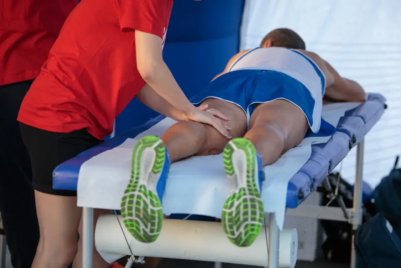 Zawodnik po zawodach otrzymuje masaż sportowy ud. Widać jego nogi w zielonych butach biegowych.