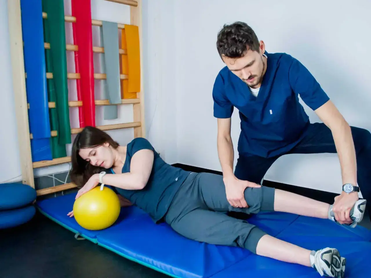 Fizjoterapeuta pomaga pacjentce w ćwiczeniach pnf. Kobieta leży na macie z żółtą piłką, a mężczyzna koryguje jej ruch.